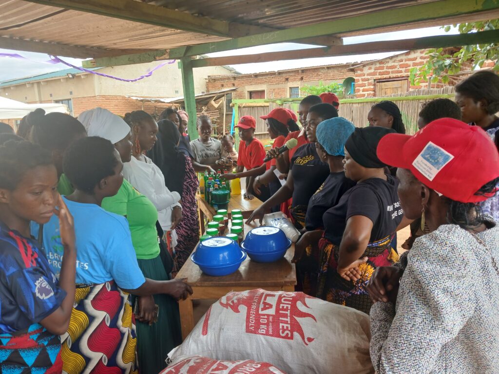 A group of women gathered around a table under a simple shelter, attentively watching a demonstration involving bowls, bottles, and containers. One woman holds a microphone while explaining the process, as others observe closely. In the foreground, large sacks and covered bowls sit on the table, suggesting a hands-on training or community workshop, likely focused on food preparation or small-scale production.
